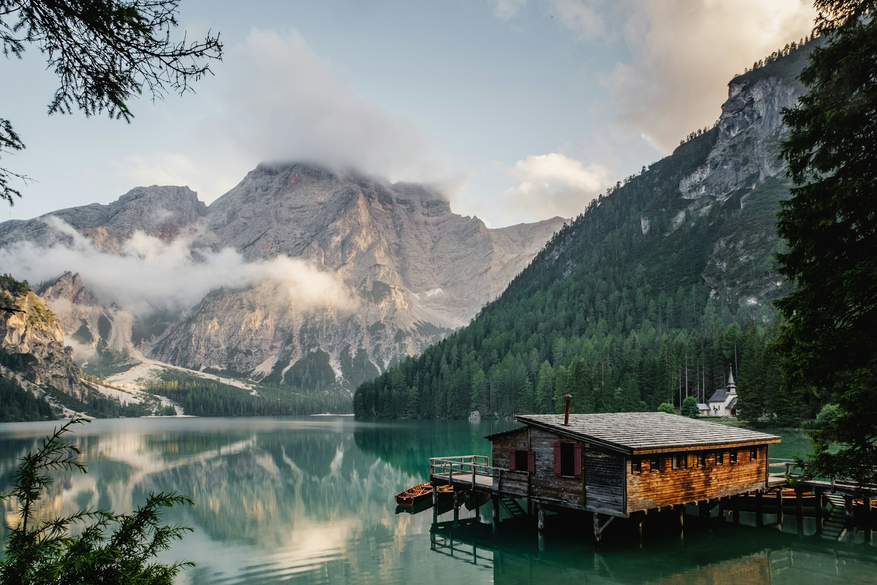 A wooden boathouse beside a still alpine lake, with forested slopes and pale mountains reflected in the water.